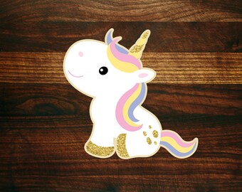 May include: A white unicorn with a pink, yellow and blue mane and tail. The unicorn has gold glitter accents and is sitting on a brown wooden surface.