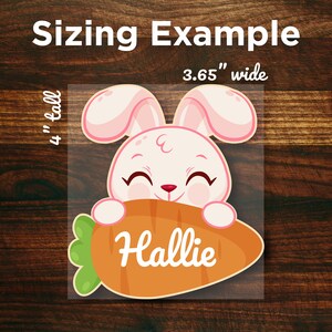 May include: A pink and white bunny rabbit with a big smile holds a carrot in its paws. The carrot is orange and green and has the name "Hallie" written on it. The image is 3.65 inches wide and 4 inches tall.
