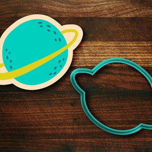 May include: A blue and yellow plastic cookie cutter shaped like a planet with a ring around it. There is a second cookie cutter shaped like a planet without a ring.