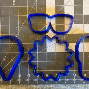 May include: Four blue plastic cookie cutters: a pair of sunglasses, a starburst, a teardrop, and a rounded rectangle.