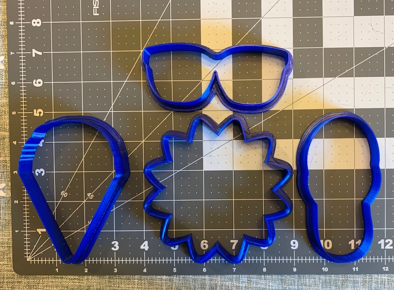 May include: Four blue plastic cookie cutters: a pair of sunglasses, a starburst, a teardrop, and a rounded rectangle.