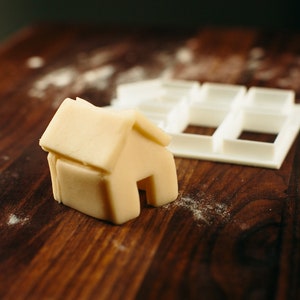 May include: A white plastic cookie cutter shaped like a house, and a small house-shaped piece of dough on a wooden surface.