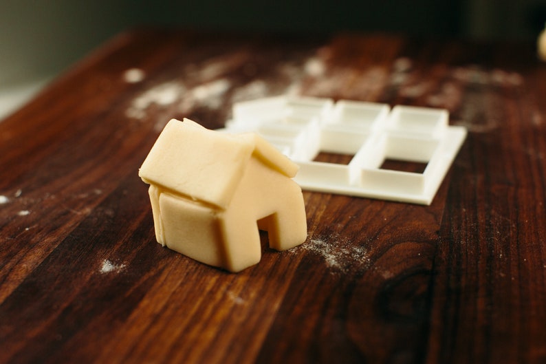 May include: A white plastic cookie cutter shaped like a house, and a small house-shaped piece of dough on a wooden surface.