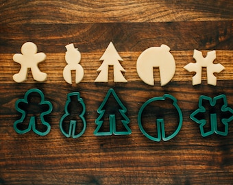 May include: Two sets of cookie cutters, one set in beige and one set in green. The beige set includes a snowman, a tree, a gingerbread person, a snowflake, and a round shape. The green set includes the same shapes.