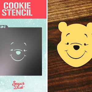 May include: A cookie stencil featuring a smiling Winnie the Pooh face. The stencil is made of plastic and is designed for use with cookie dough. The stencil is shown on a blue background with the words "Cookie Stencil" and "Sugar Dash" printed on it.