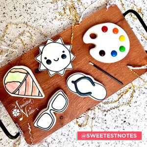 May include: A set of five decorated sugar cookies on a wooden board. The cookies are shaped like a sun, a paint palette, a pair of sunglasses, a flip-flop, and an ice cream cone. The cookies are decorated with black icing and colorful candies. The board is surrounded by gold ribbon.