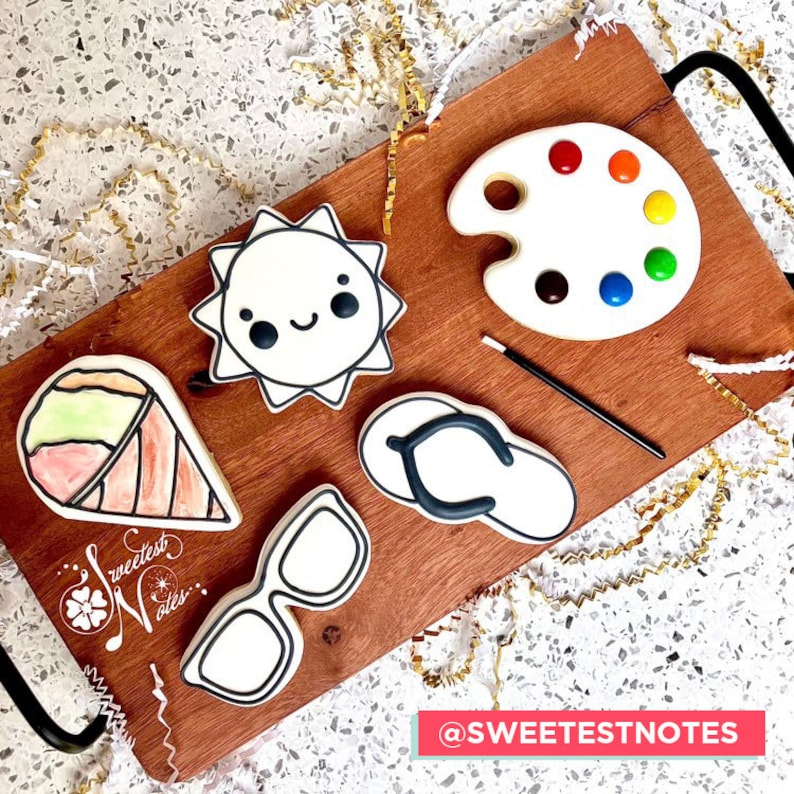 May include: A set of five decorated sugar cookies on a wooden board. The cookies are shaped like a sun, a paint palette, a pair of sunglasses, a flip-flop, and an ice cream cone. The cookies are decorated with black icing and colorful candies. The board is surrounded by gold ribbon.