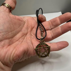 May include: A bronze-colored pentagram pendant necklace with a black cord. The pendant features a five-pointed star within a circle. A gold-colored ring with a repeating star pattern is also visible.