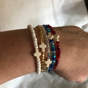 May include: A collection of beaded bracelets in various colors, each featuring a cross charm. The bracelets include white, gold, clear, blue, and red beads. The cross charms are a cream color.