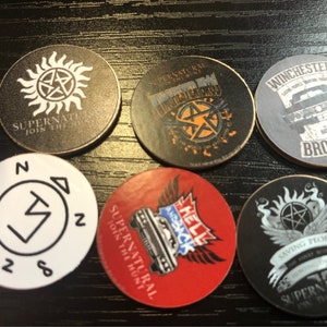 May include: Ten round, black and white, Supernatural themed, collectible tokens. The tokens feature various logos and text, including "Supernatural", "Join the Hunt", "Winchester", "Hell and Back", and "Saving People".