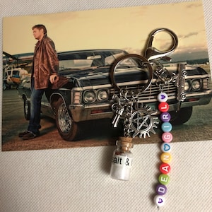May include: Keychain with a photo of a man leaning on a black car. The keychain has a bottle of salt, a pentagram charm, and colorful beads spelling "DEAN GIRL".