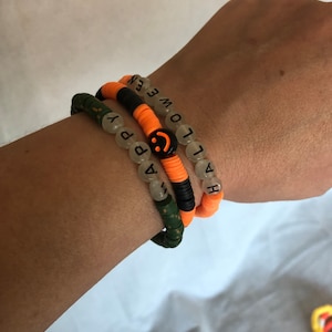 May include: Three beaded bracelets with orange and black beads. The top bracelet has the word "HALLOWEEN" spelled out in black letters. The middle bracelet has a smiley face charm and the word "HAPPY" spelled out in black letters. The bottom bracelet has green and black beads.