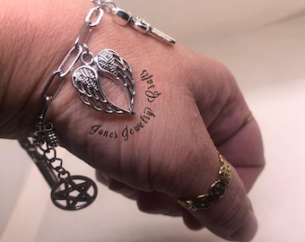 May include: A silver-toned bracelet featuring a chain link design, a key charm, and a pair of openwork angel wings. A pentagram charm hangs from the chain. The text "Jones Jewelry Crafts" is visible. A gold ring is also present.