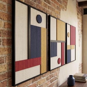 May include: A set of three rectangular wall art pieces framed in black, featuring a geometric composition of textured panels in cream, navy blue, mustard yellow, and deep red, including circular accents arranged in a structured grid pattern.