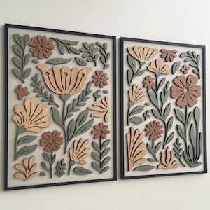 Könnte beinhalten: Zwei gerahmte florale Wandkunstwerke. Jedes zeigt ein botanisches Design mit geschichteten Holzblumen und Blättern in Braun-, Beige- und Grüntönen. Das Kunstwerk ist vor einem neutralen Hintergrund platziert und schwarz gerahmt.