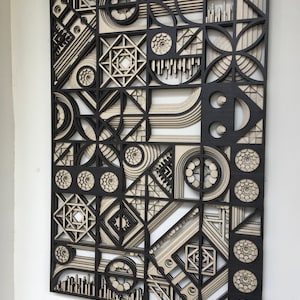 May include: A black and white geometric wall art piece made of layered wood. The design features circles, squares, and other shapes in a repeating pattern.