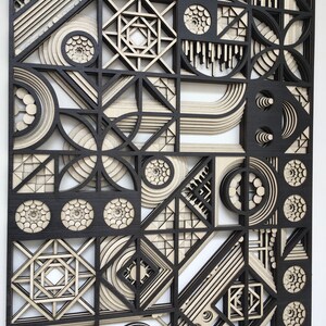 May include: A black and white geometric wall art piece made of wood. The piece features a variety of shapes, including circles, squares, and triangles. The shapes are layered on top of each other to create a three-dimensional effect.