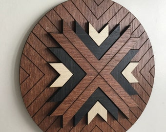 May include: A round wooden wall art piece with a geometric design. The piece features a dark brown outer ring with a chevron pattern, and a central design of layered wood in black, light brown, and cream colors.