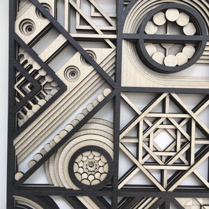 May include: A geometric wall art piece made of wood with a black and white color scheme. The art features a variety of shapes and patterns, including circles, squares, and triangles. The piece is layered and has a 3D effect.