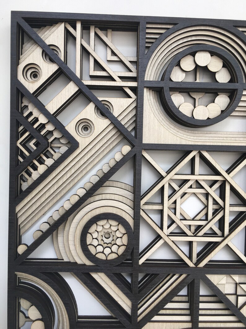 May include: A geometric wall art piece made of wood with a black and white color scheme. The art features a variety of shapes and patterns, including circles, squares, and triangles. The piece is layered and has a 3D effect.