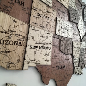 May include: A wooden map of the United States, with each state a separate piece. The map is made of different shades of brown wood, and the state names and major highways are engraved into the wood. The map is mounted on a white wall.