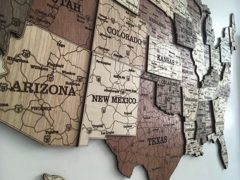 May include: A wooden map of the United States, with each state a separate piece. The map is made of different shades of brown wood, and the state names and major highways are engraved into the wood. The map is mounted on a white wall.