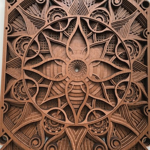 Puede incluir: Una pieza de arte de pared de madera con un intrincado dise&ntilde;o de corte l&aacute;ser que presenta un patr&oacute;n circular de mandala con un centro con forma de flor. El dise&ntilde;o est&aacute; en tonos marrones y tiene un efecto 3D.