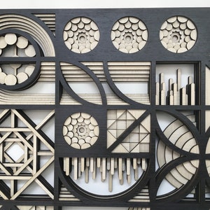 May include: A black and white geometric wall art piece with a layered design. The piece is made of wood and features a variety of shapes, including circles, squares, and triangles.