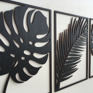 Könnte beinhalten: Drei schwarze Holz-Wanddekorationen mit ausgeschnittenen Designs von tropischen Blättern. Die linke und rechte Seite zeigen Monstera-Blätter, während die mittlere Seite eine Palmenwedel zeigt.