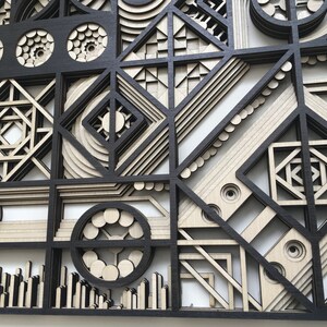 May include: A black and white geometric wall art piece made of wood. The piece is made up of various shapes and patterns, including circles, squares, and triangles. The piece is layered and has a 3D effect.
