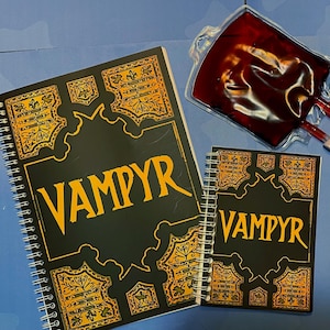 May include: Two notebooks, one spiral-bound, and a smaller notepad, both black with the word "VAMPYR" in yellow and gold decorative borders. A clear bag filled with red liquid, similar to blood, is also visible.