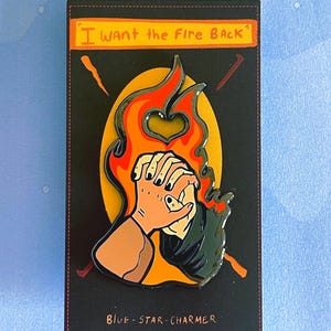 May include: Enamel pin with a design of clasped hands in front of a flame with a heart-shaped cutout. The pin is set against a black background with the text "I WANT the FIRE BACK" and "BLUE-STAR-CHARMER".