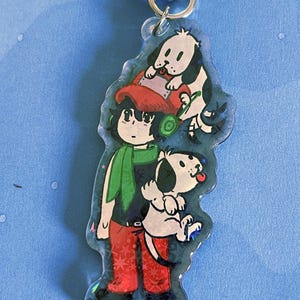 Puede incluir: Un llavero acrílico transparente con una ilustración de dibujos animados de una persona y dos perros. La persona lleva un gorro rojo, una bufanda verde y pantalones rojos. Los perros son blancos y negros. El llavero tiene una cadena de metal plateado.