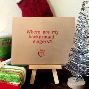 May include: A brown card with the text "Where are my background singers?!" in red lettering. A small red gift box icon is below the text. The card is on a small wooden easel.