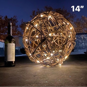 May include: A decorative sphere made of interwoven brown branches, illuminated by small, warm white lights. The sphere is approximately 14 inches in diameter. A wine bottle is next to the sphere.