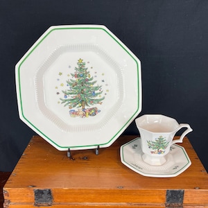May include: A white octagonal plate, teacup, and saucer set with a green trim. Each piece features a Christmas tree design with colorful ornaments and gifts. The set is displayed on a wooden box.