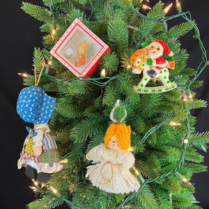 May include: A decorated Christmas tree with various ornaments. Ornaments include a red framed picture, a Santa Claus riding a rocking horse, a doll in a blue bonnet, and a yarn angel. The tree is lit with small white lights.