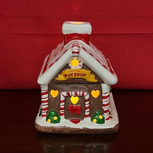 Op de afbeelding: Een keramische kerstspeelgoedwinkel met een wit dak, rood-witte strepen en een schoorsteen. De winkel heeft een bruine basis en deur, met de tekst "SANTA'S TOY SHOP" op een gele banner. Versierd met harten en kerstbomen.