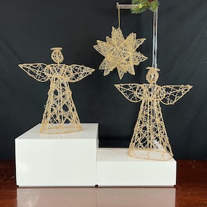 Puede incluir: Decoraciones navideñas de ángel y estrella de alambre dorado. Los ángeles tienen alas extendidas y halos. La estrella está suspendida de un soporte metálico con una ramita de vegetación y bayas rojas. Las decoraciones se exhiben sobre plataformas blancas.