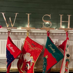 Puede incluir: Calcetines navideños festivos cuelgan de una repisa, cada uno personalizado con un nombre. Los calcetines son rojos, verdes y blancos, con diseños festivos que incluyen a Papá Noel, un árbol de Navidad y un soldado. Un letrero metálico dorado deletrea "WISH" arriba.
