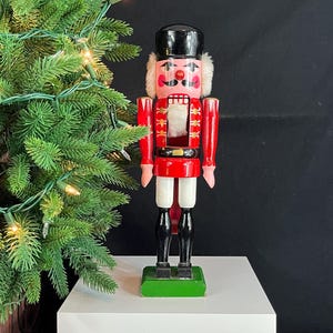 May include: A wooden nutcracker figurine in a red and white uniform with a black hat. The figurine has a pink face with white hair and is standing on a green base. A Christmas tree with lights is in the background.