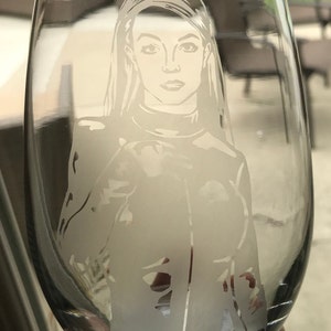 May include: A clear glass wine glass with a white etched design of a woman with long hair. The woman is wearing a collared shirt and jacket.