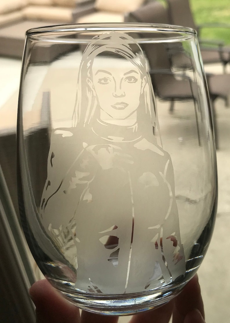 May include: A clear glass wine glass with a white etched design of a woman with long hair. The woman is wearing a collared shirt and jacket.