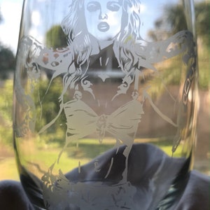 May include: A clear glass wine glass with a white etched design of a woman with long hair wearing a bikini top and a chain around her wrist.