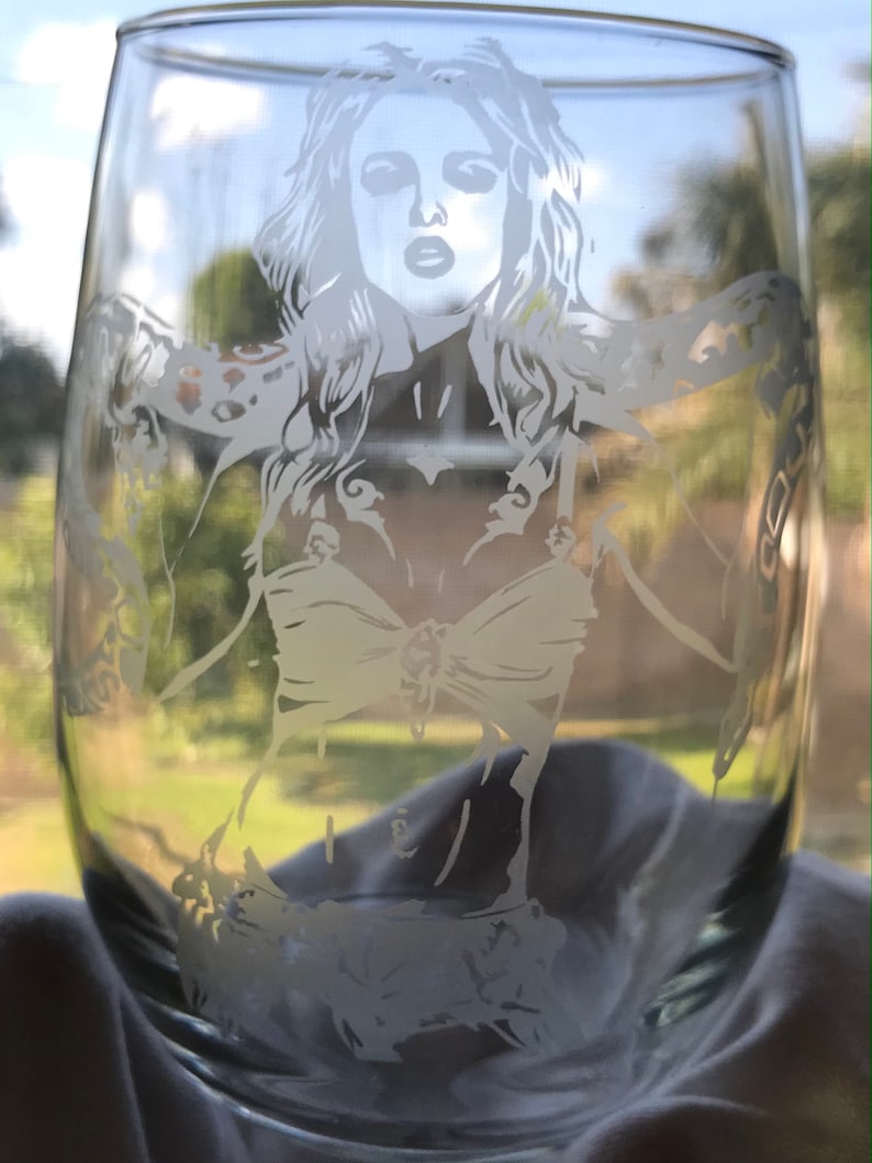 May include: A clear glass wine glass with a white etched design of a woman with long hair wearing a bikini top and a chain around her wrist.