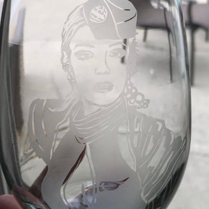 May include: A clear glass wine glass with a frosted etching of a woman wearing a hat and a scarf. The etching is detailed and shows the woman's face, hair, and clothing.