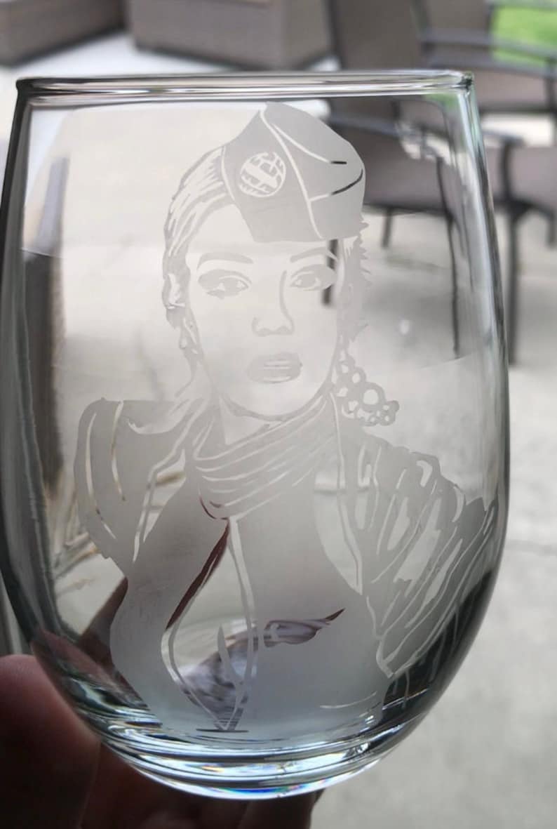 May include: A clear glass wine glass with a frosted etching of a woman wearing a hat and a scarf. The etching is detailed and shows the woman's face, hair, and clothing.