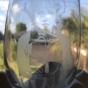 May include: A clear glass wine glass with a white etched design of a woman with a hairstyle similar to Princess Leia from Star Wars.