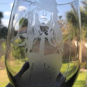 May include: A clear glass wine glass with a frosted etching of a woman wearing a hat and a collared shirt.