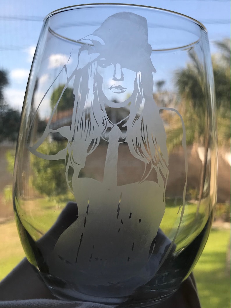 May include: A clear glass wine glass with a frosted etching of a woman wearing a hat and a collared shirt.
